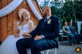 In Budapest, Hungary, at the ceremony location, the bride and groom sit in chairs outside at the altar, holding hands and sharing a laugh, highlighting their joyful connection.